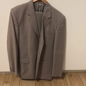 Grå kostym med kavaj och byxor - Snygg grå kostym med klassisk kavaj och matchande kostymbyxor. Kavajen har breda slag, två framfickor och knappar vid ärmslut. Byxorna har pressveck, bälteshällor och stängs med dragkedja och knapp. Perfekt för dig som vill ha en stilren och tidlös look.