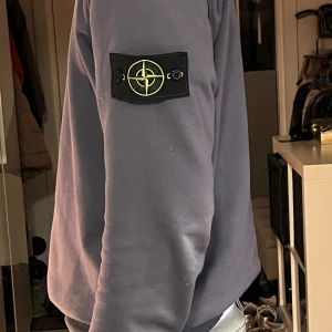 Blå sweatshirt från Stone Island - Säljer en grå sweatshirt från Stone Island med den klassiska logotypen på vänster ärm. Tröjan har rund hals, ribbade muddar och en clean look som är enkel att matcha. Perfekt för dig som gillar streetwear och vill ha något stilrent.