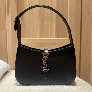 Svart axelväska från Saint Laurent - Snygg svart axelväska från Saint Laurent i slät skinn med ikoniskt YSL-emblem i guld på framsidan. Väskan har en justerbar rem och dragkedja upptill. Perfekt storlek för det viktigaste och ger en lyxig vibe till din outfit. Första bild lånad men samma väska😊💕