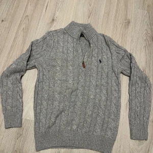Grå stickad tröja Polo Ralph Lauren - Snygg grå stickad tröja från Polo Ralph Lauren med klassiskt kabelmönster och halv dragkedja vid kragen. Tröjan har långärmade ribbade muddar och en broderad marinblå logga på bröstet. Perfekt för dig som gillar stilren och tidlös design.