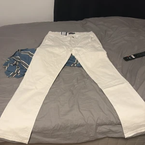 Vita chinos från Dressmann slim fit - Snygga vita chinos från Dressmann i slim fit-modell. Byxorna har klassiska fickor fram och bak med knappdetaljer, bälteshällor och är tillverkade i stretchig bomull för extra komfort. Perfekt för dig som gillar stilrena och fräscha byxor. Helt nya och kvitto finns och alla lappar sitter på.