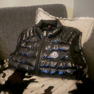 Svart dunväst från Moncler, S - Svart, glansig dunväst från Moncler med quiltad design och dragkedja framtill. Västen har hög krage och Moncler-logga på bröstet samt logotypband längs axlarna. Perfekt för lager-på-lager och streetstyle. Snygg, sportig look med normal passform. INTE RIKTIGT!!!!