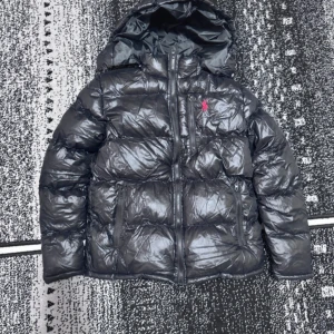 Svart pufferjacka från RL - Svart pufferjacka från RL med glansig finish och röd broderad logga på bröstet. Jackan har huva, två sidofickor med dragkedja och quiltad design. Perfekt för kalla dagar och har en riktigt chill vibe.