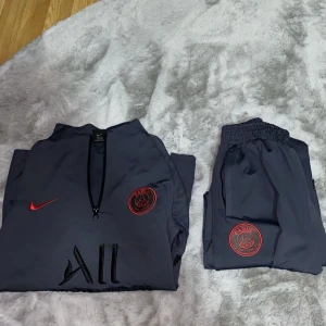 PSG träningsset Nike mörkgrå - Säljer ett fett PSG träningsset från Nike i mörkgrått med röda detaljer. Tröjan har lång ärm, halv dragkedja och Paris Saint-Germain-logga på bröstet. Byxorna har elastisk midja, PSG-märke och Nike Swoosh i rött. Materialet är lätt och andas, perfekt för träning. Storlek m men passar s 