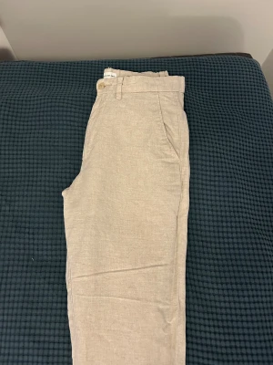  Beige byxor från These Glory Days - Snygga beigebyxor från These Glory Days med klassisk rak passform och diskreta fickor både fram och bak. Byxorna har bälteshällor och stängs med knapp och dragkedja. Perfekta för en chill och stilren look.