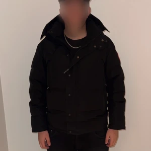Svart dunjacka från Canada Goose - Svart dunjacka från Canada Goose med klassisk patch på bröstet och hög krage. Jackan har huva och knäppning med både dragkedja och knappar. Perfekt för kalla dagar och riktigt snygg streetsti. Jackan sitter som Large i slim. Passar dig som Medium regular ish om man är ungefär 180-190 cm. Can också byta mot en Canada goose macmillan I L-XL svart