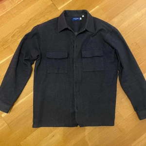 Jack and Jones overshirt mörkblå  - Snygg mörkblå overshirt från Jack and Jones med klassisk krage och två stora bröstfickor med lock. Skjortan har långa ärmar och knäppning framtill. Materialet är mjukt och känns som ull, perfekt för lager på lager. Enkel och stilren design som funkar till många outfits. Storlek S