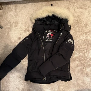 Svart dunjacka Moose Knuckles med päls - Svart dunjacka från Moose Knuckles med fluffig vit päls runt huvan, coolt broderi på insidan och metallpatch på ärmen. Jackan har dragkedja, knappar och flera fickor. Perfekt för kalla dagar och har en riktigt snygg streetstil.