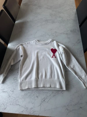 Vit stickad tröja från Ami - Vit stickad tröja från Ami med röd hjärtbroderi och bokstaven A på bröstet. Tröjan har rund hals, ribbade muddar och nederkant samt långärmad passform. Perfekt för dig som gillar stilrena detaljer och vill ha något som sticker ut lite extra.