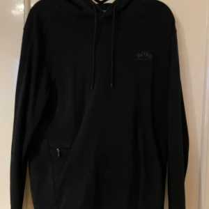 Svart hoodie från BOSS med huva - Svart hoodie från BOSS med diskret logga på bröstet, huva med snörning och långa ärmar. Varan är använd i bra skick utan skador. Den säljs i befintligt skick. 
