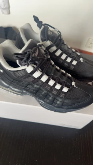 Nike Air Max 95 svart/vit sneakers - Nike Air Max 95 sneakers i svart och vitt med ikonisk vågig design och synlig Air-dämpning i sulan. Ovandel i mesh och skinn med vita detaljer och snörning. Perfekta för dig som vill ha en klassisk och sportig look med maximal komfort.