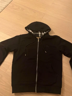 Svart Burberry hoodie med dragkedja - Svart hoodie från Burberry med klassiskt rutigt foder i huvan. Tröjan har dragkedja framtill, snörning vid huvan och två stora fickor. Materialet är mjuk bomull och passformen är normal. Perfekt för dig som gillar stilrena och lyxiga streetwear-plagg.