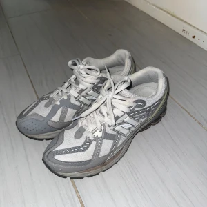 New Balance 1906  - Säljer ett par New Balance 1906 sneakers i grått och vitt med snygga detaljer och logga på sidan. Skorna har snörning, mesh och syntetpaneler för bra ventilation och komfort. Sulan är stabil och har en cool design baktill. Perfekt för dig som gillar retrostil och streetwear.