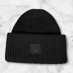 Svart ribbstickad mössa från Acne Studios - Svart ribbstickad mössa från Acne Studios med klassisk uppvikt kant och den ikoniska ansiktsloggan framtill. Tjockt stickad i mjukt material som håller dig varm och ger en clean look. Perfekt för dig som gillar minimalistisk stil.