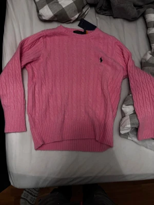 Rosa kabelstickad tröja Polo Ralph Lauren - Snygg rosa kabelstickad tröja från Polo Ralph Lauren med klassisk rund hals och broderad logga på bröstet. Tröjan har långa ärmar och breda ribbade muddar vid ärmslut och nederkant. Perfekt för dig som gillar preppy stil och vill ha något mjukt och bekvämt.