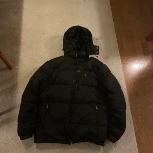 Svart pufferjacka från Polo Ralph Lauren - Svart pufferjacka från Polo Ralph Lauren med röd broderad logga på bröstet. Jackan har huva, flera dragkedjeförsedda fickor och en quiltad design. Perfekt för kalla dagar och har en modern, sportig vibe.