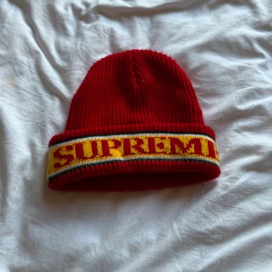 Röd Supreme mössa med gul kant - Supreme mössa i rött med bred uppvikt kant i gult, svart och vitt. Supreme-loggan är stickad i gult på kanten. Ribbstickad och tillverkad i 100% akryl, perfekt för att sticka ut i vinterkylan.