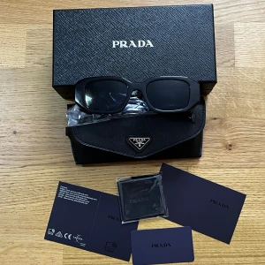  Prada Solglasögon  - Svarta solglasögon från Prada med rektangulär form och breda skalmar. Snyggt Prada Milano-tryck på sidan och tillverkade i Italien. Kommer med originalfodral, putsduk och certifikat. Perfekt accessoar för dig som gillar stilren och exklusiv design.