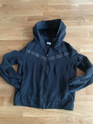 Svart Nike tech fleece med dragkedja - Svart hoodie från Nike med hel dragkedja och huva. Snygga gråa ränder över bröstet och ärmen, samt diskret Nike-logga på vänster sida. Jackan har lång ärm och är gjord i mjuk bomull, perfekt för chill eller träning.