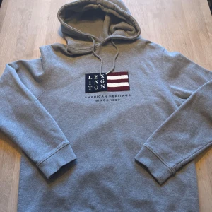 Grå Lexington hoodie med broderi - Snygg grå hoodie från Lexington i storlek L. Tröjan har huva med snörning och ett broderat Lexington-märke på bröstet med marinblå och vinröda detaljer. Mjuk insida och ribbade muddar vid ärmslut och nederkant. Perfekt för chill dagar.