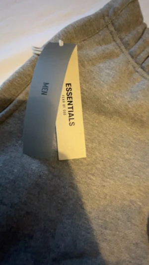 Grå Essentials mjukisbyxor med snörning - Grå mjukisbyxor från Essentials Fear of God med elastisk midja och långa snören framtill. Byxorna har ribbade muddar vid bensluten och logotyptryck på vänster ben. Perfekt för chill och streetwear. Materialet är mjukt och bekvämt bomull.