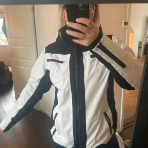 Vit och svart vindjacka med huva - Snygg vit och svart vindjacka med dragkedjor på bröstet och sidorna. Jackan har huva, svarta detaljer längs ärmar och sidor samt en sportig look. 