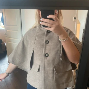Beige kappa med vida ärmar och knappar - Säljer en beige cape med vida, korta ärmar och stora knappar framtill. Jackan har två stora fickor och en snygg, rak passform. 