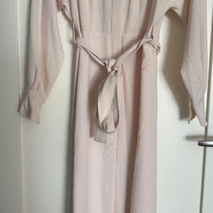 Ljusbeige långklänning  - En stilren och enkel långklänning i ljusbeige ton från Mango med långa ärmar och knytbart bälte i midjan. Klänningen har rak passform och är tillverkad i ett mjukt, slätt tyg som faller snyggt. Perfekt för dig som gillar minimalistisk och clean stil.