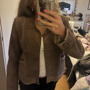 Brun mockajacka med knappar - Säljer en snygg brun lite vintage mockajacka med klassisk krage och knäppning framtill. Jackan har två fickor och en diskret rutig insida vid ärmslutet. Perfekt för dig som gillar stilrena och tidlösa plagg med lite edge. Storlek 40 men skulle säga att den passar m och s.