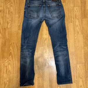 Nudie jeans - Säljer ett par blå jeans med raka ben och snygga slitningar. Jeansen har klassiska bakfickor och kontrastsömmar. Perfekta för en avslappnad och trendig look.