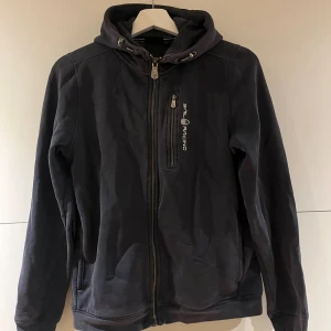 Sail Racing hoodie med dragkedja - Två snygga hoodies från Sail Racing, en i marinblå och en i vitt. Båda har huva med snörning, hel dragkedja och en bröstficka med Sail Racing-logga. Perfekt för dig som gillar sportig och clean stil. Materialet är mjuk bomull och polyesterblandning.