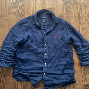 Mörkblå linneskjorta från Polo Ralph Lauren - Snygg mörkblå skjorta från Polo Ralph Lauren i linne med klassisk krage och knappar framtill. Ikonisk röd broderad logga på bröstet. Perfekt för en stilren och avslappnad look. Inga defekter har en exakt likadan fast vit 