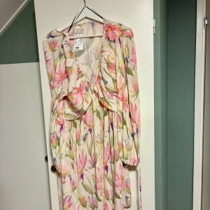 Blommig kortklänning från H&M - Superfin kortklänning från H&M med blommigt mönster i rosa, grönt och blått på ljus botten. Klänningen har v-ringad hals, volangdetaljer och långa ärmar med snörning vid ärmslut. Materialet är plisserad polyester som ger en luftig och flowy känsla.