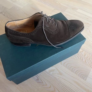 Bruna mocka westbourne från Crockett & Jones - Snygga Westbourne skor från Crockett & Jones i mörkbrun mocka med klassisk snörning och dekorativa broguemönster. Skorna har en spetsig tå och platt sula, perfekt för dig som gillar stilrena och tidlösa detaljer. Levereras med originalkartong och dustbags. Orginalpris 7864 kronor. 