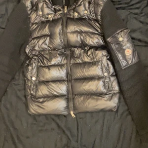 Svart pufferjacka från Moncler - Säljer en svart pufferjacka från Moncler med glansig yta och stickade ärmar. Jackan har dragkedja framtill, tryckknappar på fickorna och Moncler-logga på ärmen. Perfekt för dig som vill ha en snygg och varm jacka med coola detaljer. 