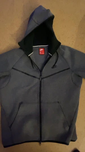 Grå hoodie från Nike med dragkedja - Snygg grå hoodie från Nike i storlek M med svart Nike-logga på bröstet. Tröjan har hel dragkedja, huva med snörning och två fickor framtill. Materialet är mjukt och känns som en blandning av bomull och polyester, perfekt för chill eller träning.