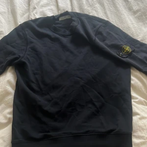 Mörkblå sweatshirt från Stone Island - Mörkblå sweatshirt från Stone Island med klassisk rund hals och ikonisk patch med logga på vänster ärm. Tröjan har en clean look och är tillverkad i mjuk bomull som känns skön mot huden. Perfekt för dig som gillar streetwear och stilrena detaljer.