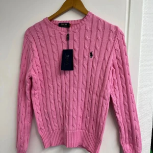 Polo Ralph Lauren Tröja - Riktigt snygg rosa kabelstickad tröja från Polo Ralph Lauren med klassisk rund hals och broderad logga på bröstet. Tröjan har långa ärmar och ribbade muddar vid ärmslut och nederkant. Perfekt för dig som gillar preppy stil och vill ha något som sticker ut.