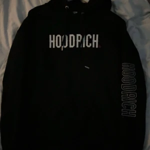 Svart hoodie från Hoodrich XS - Svart hoodie från Hoodrich i storlek XS med stor vit logga på bröstet och ärmen. Baksidan har tryck med text, röd detalj och grafiska element. Hoodien har huva med snörning och är långärmad.