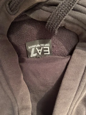Svart hoodie från Emporio Armani EA7 - Svart hoodie från Emporio Armani EA7 med stort logotryck i grått och guld på bröstet. Tröjan har huva med snörning och en klassisk magficka. Mjuk insida och ribbade muddar vid ärmslut och nederkant. Perfekt för dig som gillar stilrena märkesplagg.