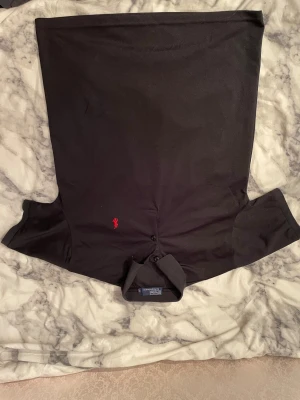 Svart pikétröja från Polo Ralph Lauren - Svart pikétröja från Polo Ralph Lauren med klassisk krage och två knappar framtill. Tröjan har ett rött broderat logomärke på bröstet och är tillverkad i mjuk bomull. Perfekt för dig som gillar stilrena och tidlösa plagg.