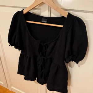 Svart blus med puffärm från Gina Tricot - Supersöt svart blus från Gina Tricot i storlek S. Blusen har puffiga korta ärmar, ribbad struktur och två knytband framtill. Modellen är lite croppad med volang nedtill och har en rundad halsringning. Den vita toppen är bara för att visa hur den ser ut på.