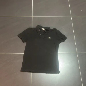 Svart pikétröja Lacoste slim fit M - Svart pikétröja från Lacoste i slim fit-modell, storlek M. Klassisk design med krage, korta ärmar och den ikoniska gröna krokodilloggan på bröstet. Tröjan är gjord i mjuk bomull och har knappar vid halsen för en clean look. Den passar även en stor S