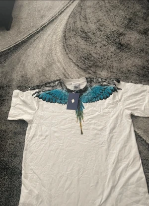Vit t-shirt med blå vingar Marcelo Burlon - Säljer en vit t-shirt från Marcelo Burlon County of Milan i storlek L. T-shirten har ett fett tryck med blå och svarta vingar runt halsen och är gjord i mjuk bomull. Klassisk passform och korta ärmar, perfekt för dig som gillar statement-plagg.