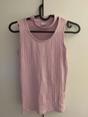 Ljusrosa ribbad linnetopp Gina Tricot - Supersöt ljusrosa linnetopp från Gina Tricot i ribbad, mjuk viskosblandning. Toppen är ärmlös med rund hals och croppad passform som sitter snyggt mot kroppen. Perfekt att styla med jeans eller shorts för en chill look.