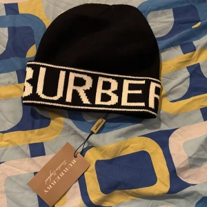 Svart Burberry mössa  - Svart mössa från Burberry med bred vit kant där Burberrys logga är stickad i stora bokstäver. Klassisk och stilren design som ger en lyxig vibe. Materialet är stickat och känns mjukt och bekvämt. Perfekt för dig som vill ha något snyggt och trendigt på huvudet.