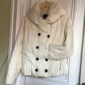 Vit pufferjacka från Kriss - Säljer en vit pufferjacka från Kriss med stor krage och dubbelknäppning med svarta knappar. Jackan har lång ärm och två sidofickor med dragkedja. Perfekt för dig som vill ha en varm och snygg jacka med cool siluett.