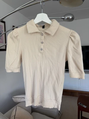 Beige ribbad blus från Pieces XS - Säljer en beige ribbad blus från Pieces i storlek XS. Toppen har puffade korta ärmar, klassisk krage och fyra dekorativa knappar framtill med glittriga detaljer. Tillverkad i mjuk ekologisk bomull med lite stretch för skön passform.