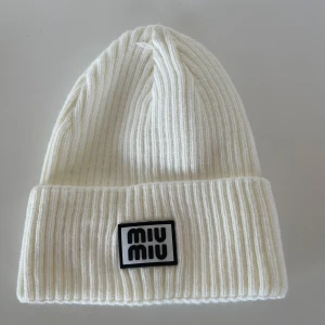 Vit ribbad mössa från Miu - Snygg vit mössa från Miu  med ribbad struktur och uppvikt kant. Framtill sitter en svartvit logotyp-patch som ger en cool touch. Perfekt för dig som vill ha en stilren och trendig accessoar till vintern.
