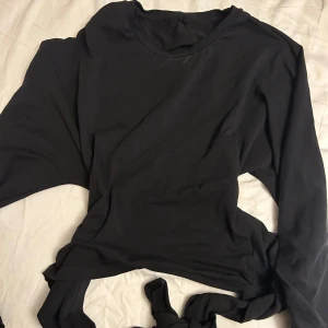 Svart långärmad croppad tröja - Svart långärmad tröja från Get To Know Your Garment med rund hals och croppad passform. Tröjan har en snygg knyt-detalj i ryggen som gör att du får en snygg passform och som ger en cool vibe. Tillverkad i mjuk bomull som känns skön mot huden. Perfekt för dig som gillar stilrena och enkla plagg.